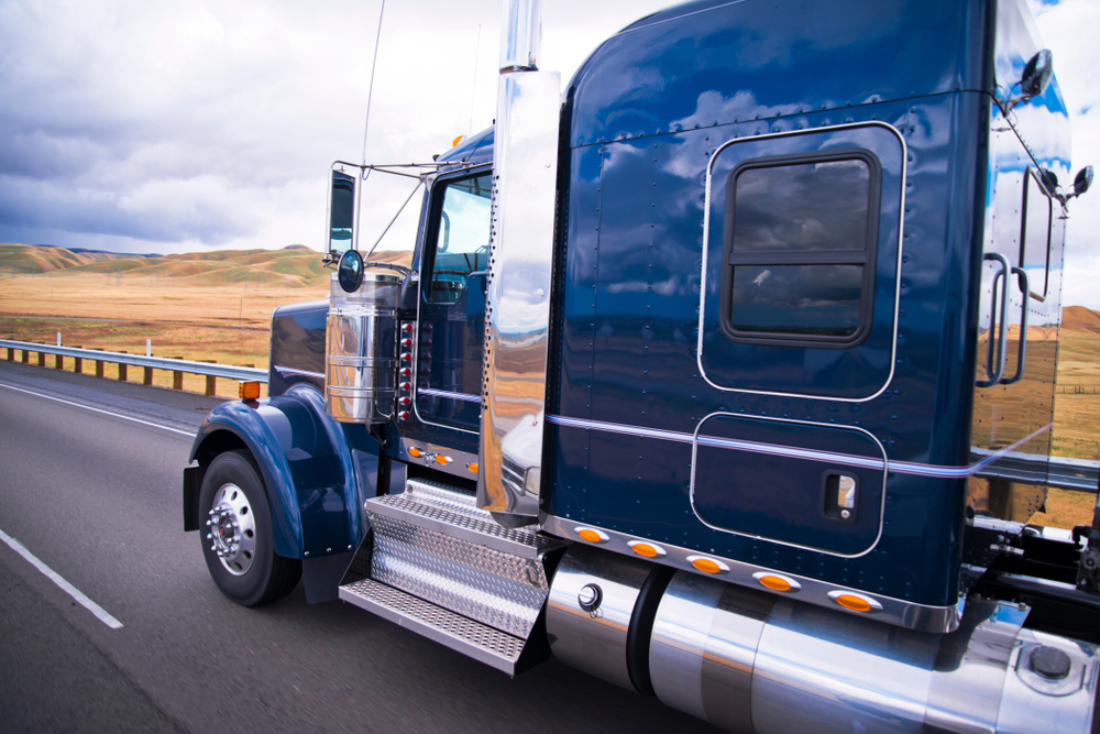 A dark navy blue heavy truck cab drives down a highway. The surrounding blue sky with clouds and the fields are reflected on the side.