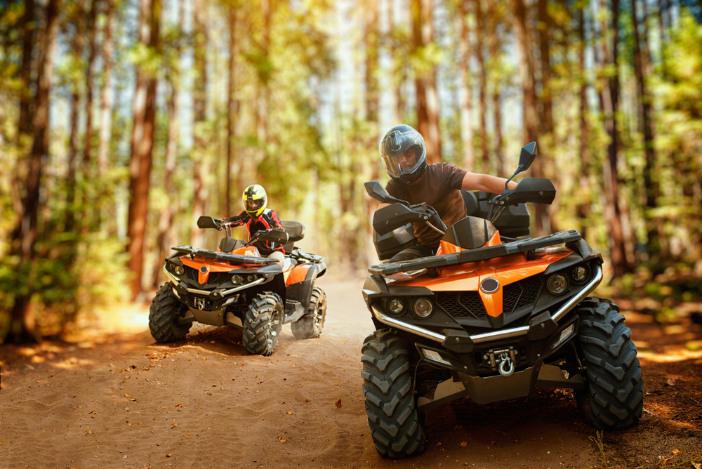 Two ATVs move through rough terrain surrounded by trees.