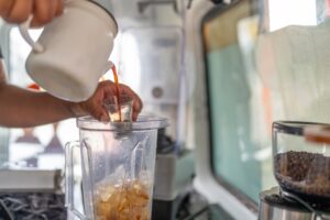 A barista pours a shot of espresso into a blender for a frozen drink in a busy cafe environment.