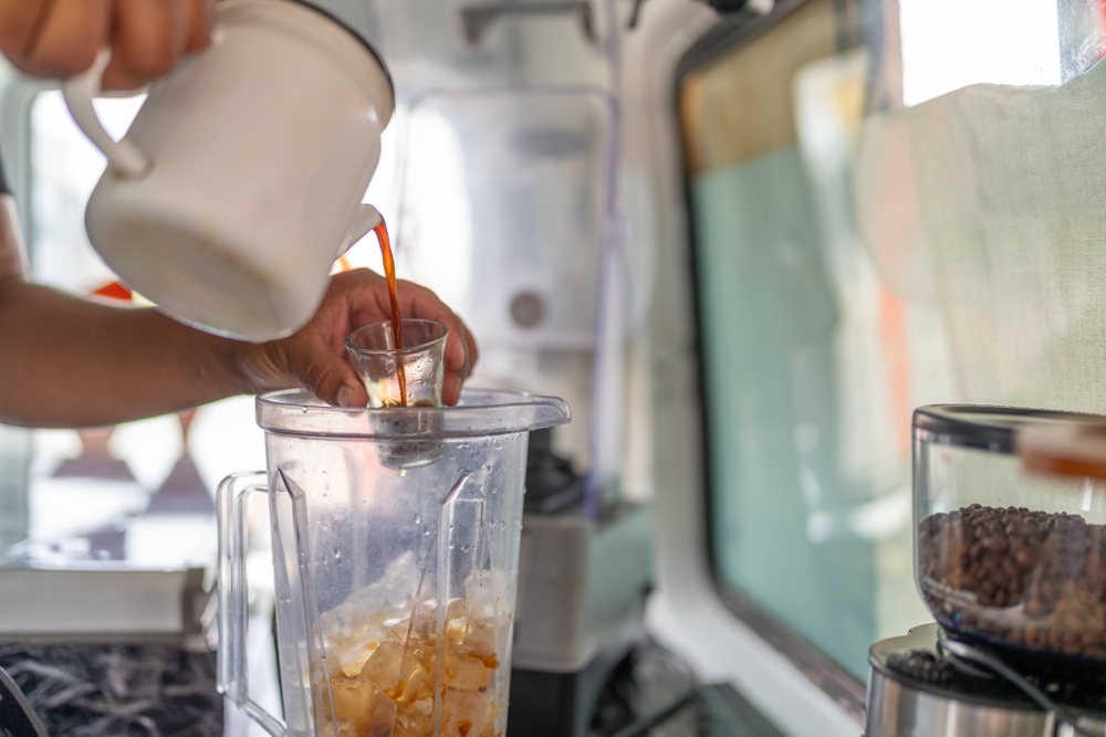 A barista pours a shot of espresso into a blender for a frozen drink in a busy cafe environment.